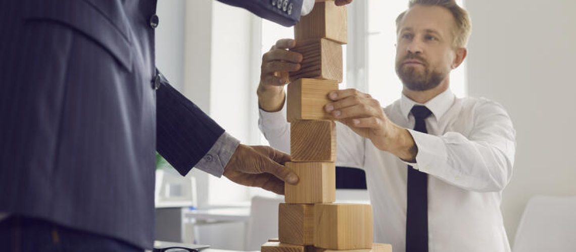 Mature and young businessmen making wood block tower as metaphor for building strong business together. Concepts of collaboration, succession, maintaining financial stability, economical growth